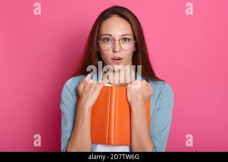 Horizontal shot of cute emotional impressed young female opening eyes and mouth widely, holding book in both hands, standing isolated over pink backgr Stock Photo