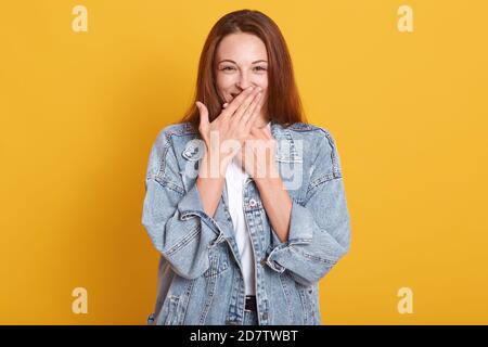Happy good looking young model covering her mouth with both hands, having shy surprised look, being in high spirits, wearing denim jacket and white t Stock Photo