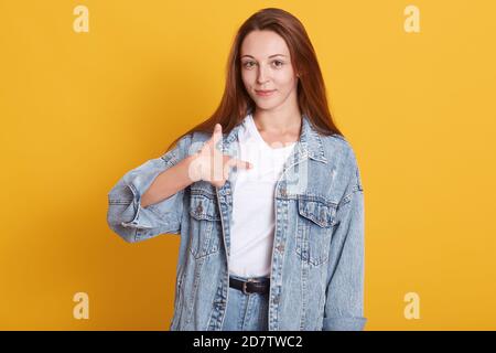 Close up portrait of pretty young woman with long dark staright hair wearing denim jacket and white shirt, standing and pointing at herself isolated o Stock Photo