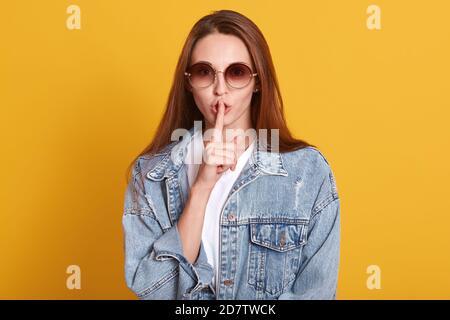 Portrait of mysterious stylish pretty model making gesture, putting her forefinger to lips, looking directly at camera, keeping secrets, wearing white Stock Photo