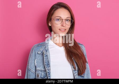 Indoor shot of pleasant looking woman, smiles positively, wearing optical glasses, white t shirt and denim jacket, being in hight spirt, model posing Stock Photo