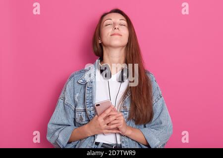 Portrait of young girl wearing stylish denim jacket, holding mobile phone in hands, keeps head up with closed eyes, fells grateful, glad to recive mes Stock Photo