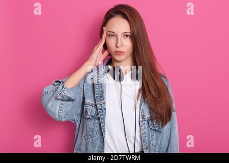 Indoor shot of young woman in denim casual clothes, holding headphones around neck, has headache from loud music, looks tired, keeping finger on templ Stock Photo
