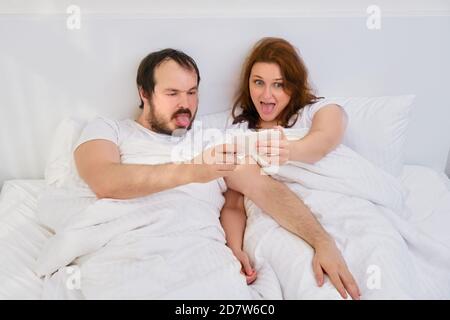 A man and a woman take a selfie photo funny in a white bed. Husband and wife spend time at home under the coronavirus quarantine. Stock Photo