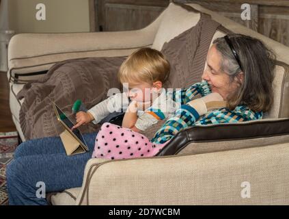 Grandmother helping small baby boy with app on a digital tablet while sitting in family room and relaxing Stock Photo