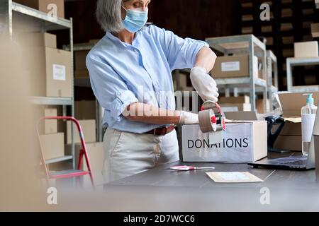 Mature female warehouse worker wearing mask packing donations box. Stock Photo