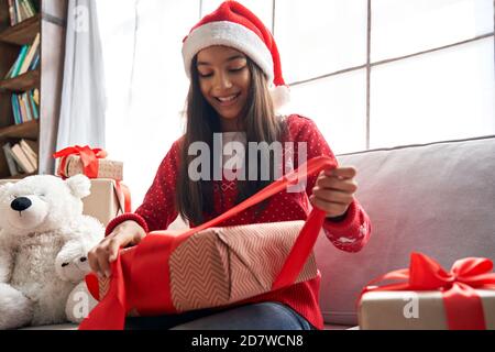 Happy indian latin kid girl unties red ribbon opens Christmas gift box at home. Stock Photo