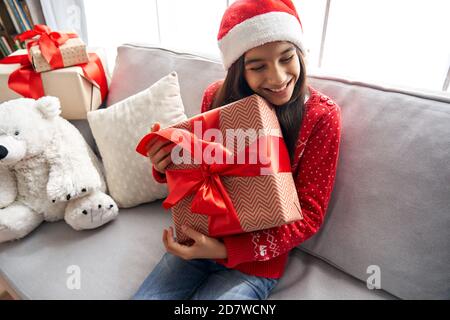 Happy cute indian kid girl holding gift box Christmas present sitting on couch. Stock Photo