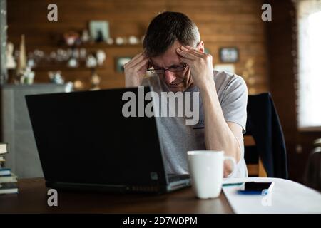 Frustrated mid adult man touching his head and keeping eyes closed while sitting in front of laptop. Stock Photo