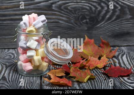 Marshmallows of different colors. They lie in a glass jar. Autumn maple leaves are scattered around. Stock Photo