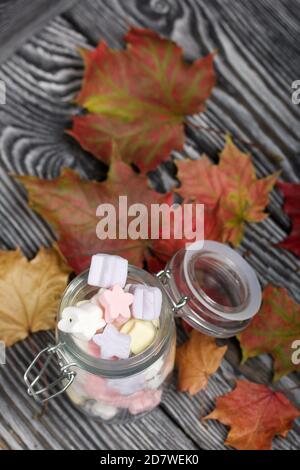 Marshmallows of different colors. They lie in a glass jar. Autumn maple leaves are scattered around. Stock Photo