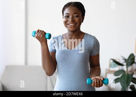 Domestic Training. Cheerful sporty african american woman exercising with dumbbells at home Stock Photo