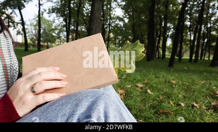 A woman holds a closed book lying in her lap with a fallen oak leaf close-up in a park on a sunny warm autumn day. The concept of relaxation, reading Stock Photo