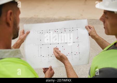 Back view at two building contractors looking at plans while standing at construction site, focus on floor plans Stock Photo