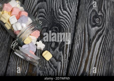 Marshmallows of different colors. They lie in an overturned glass jar. Against the background of black pine boards. Stock Photo
