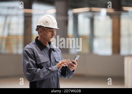 Side view portrait of mature worker using smartphone while standing at construction site or in industrial workshop, copy space Stock Photo