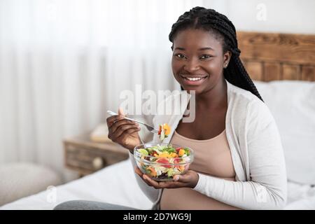 Black Expectant Woman Eating Fresh Vegetable Salad, Sitting On Bed At Home Stock Photo