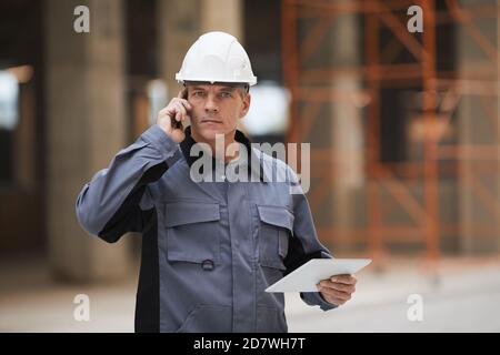 Waist up portrait of mature worker speaking by smartphone and looking at camera while standing at construction site or in industrial workshop, copy space Stock Photo