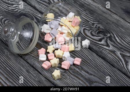 Marshmallows of different colors. They lie in an overturned glass jar. Against the background of black pine boards. Stock Photo