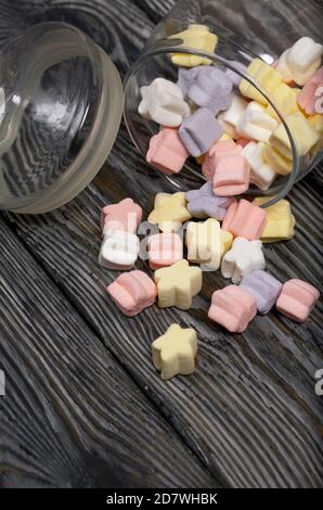 Marshmallows of different colors. They lie in an overturned glass jar. Against the background of black pine boards. Stock Photo