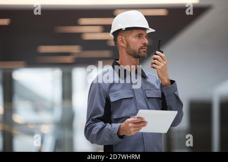 Waist up portrait of handsome mature worker speaking by walkie-talkie while supervising work at construction site or in industrial workshop, copy space Stock Photo