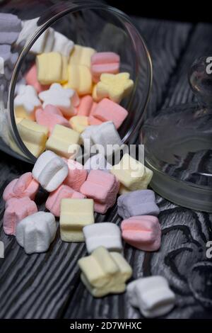 Marshmallows of different colors. They lie in an overturned glass jar. Against the background of black pine boards. Stock Photo