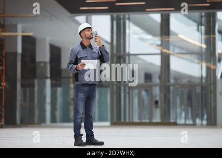 Full length portrait of mature worker speaking by walkie-talkie while supervising work at construction site or in industrial workshop, copy space Stock Photo