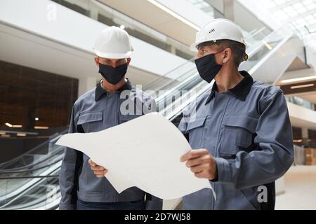 Waist up portrait of two construction workers wearing masks and discussing plans while standing in shopping mall or office building Stock Photo