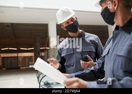 Waist up portrait of two construction workers wearing masks and discussing plans while standing in shopping mall or office building, copy space Stock Photo
