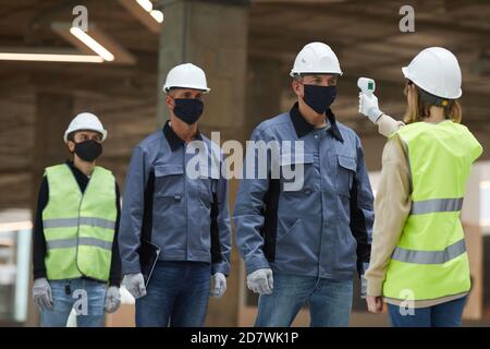 Portrait of female supervisor measuring temperature of workers with contactless thermometer at construction site, corona virus safety measures Stock Photo