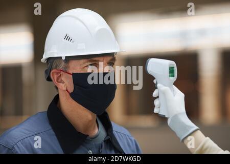 Portrait of mature worker wearing mask and waiting to measure temperature with contactless thermometer at construction site, corona virus safety Stock Photo