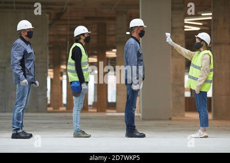 Side view full length of female supervisor measuring temperature of workers with contactless thermometer at construction site, corona virus safety measures Stock Photo