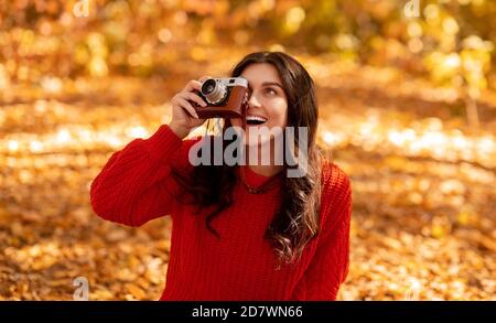 Smiling young woman in bright red sweater making pictures with retro camera at park on sunny fall day Stock Photo