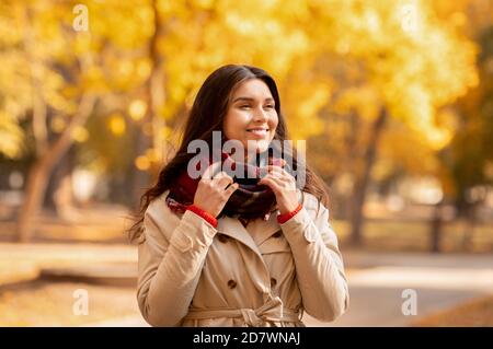 Beautiful millennial lady in coat and warm scarf enjoying colorful change of seasons in autumn park Stock Photo