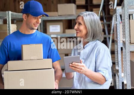 Happy old female supervisor using tablet talking to male courier holding boxes. Stock Photo