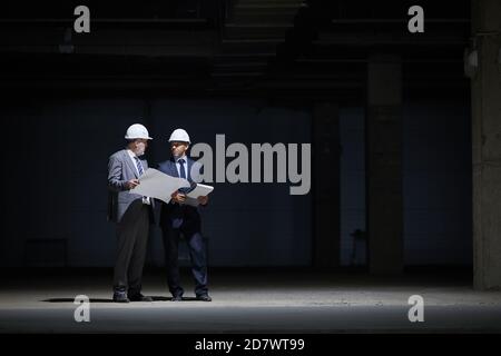 Dramatic full length portrait of two mature business people wearing hardhats and holding plans while standing in dark at construction site lit by harsh lighting, copy space Stock Photo