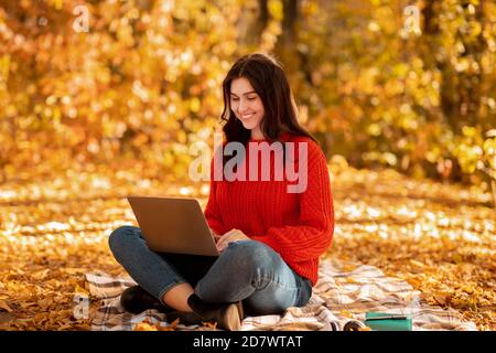 Lovely Caucasian woman using laptop for online work, browsing web at city park in autumn Stock Photo