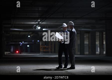 Dramatic full length portrait of two business people wearing hardhats and holding plans while standing in dark at construction site, copy space Stock Photo