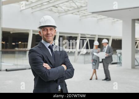 Waist up portrait of smiling mature businessman wearing hardhat and looking at camera while standing with arms crossed at construction site, copy space Stock Photo