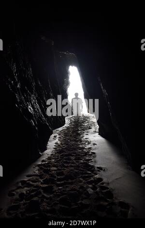 A transparent ghostly figure walks towards the light at the end of a tunnel, heading out of a cave with sandy footprints leading out from the front. Stock Photo