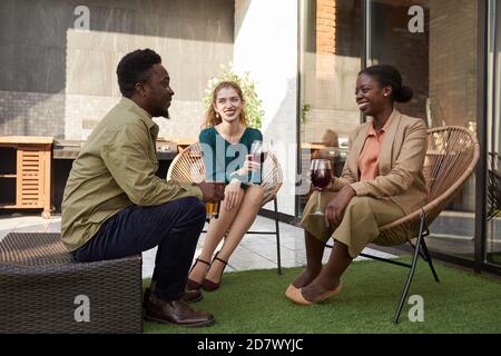 Full length portrait of contemporary adult people enjoying conversation while sitting on wicker chairs during outdoor party Stock Photo