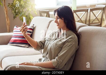 Smiling young indian woman sit on couch using mobile phone video call. Stock Photo