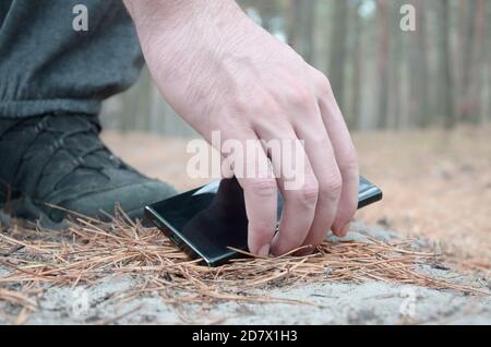 Male hand picking up a lost mobile phone from a ground in autumn fir wood path. The concept of finding a valuable thing and good luck Stock Photo