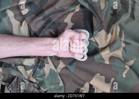 Male fist with brass knuckles on the background of a camouflage jacket. The concept of skinhead culture, homemade melee weapons Stock Photo