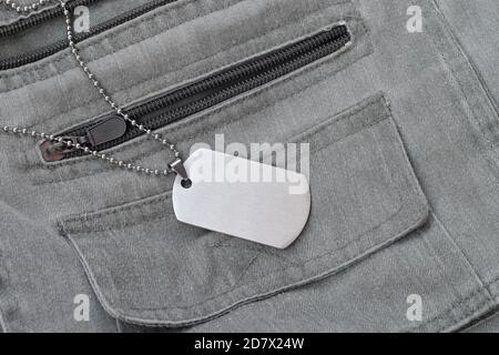 Silvery military beads with dog tag on dark grey vest with pockets. Army token on soldiers camo jacket Stock Photo