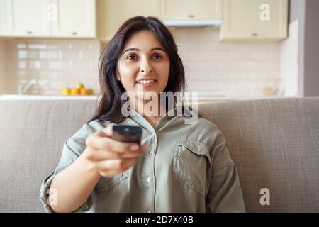 Happy indian woman sitting on sofa holding remote control watching tv at home. Stock Photo