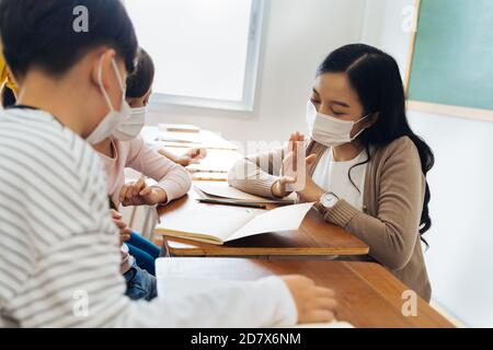 Close-up of Asian female teacher wearing a face mask in school building tutoring a primary student children. Elementary pupils writing and learning in classroom. Covid-19 school reopen concept Stock Photo