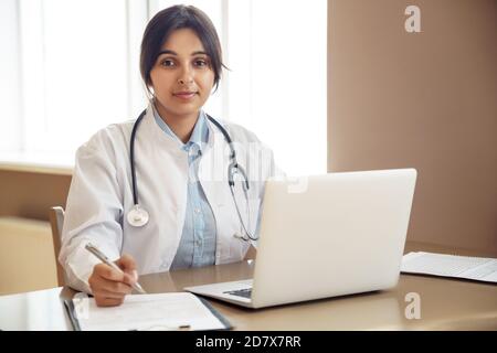 Young indian female doctor wear white coat looking at camera at workplace. Stock Photo