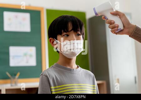Asian male student in school building checked and scanned for temperature check. Elementary pupil wearing a face mask and line up before entering into classroom. Covid-19 school reopen concept Stock Photo