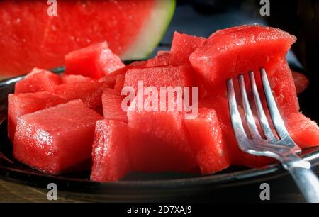 A fork with a cube of watermelon over some more watermelon cubes on a black plate and a watermelon slice and a whole watermelon in the background Stock Photo